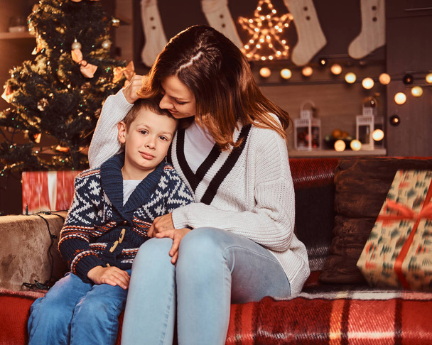 Mom hugging her cute little boy while sitting on sofa in decorated room during Christmas time. - Foto, afbeelding