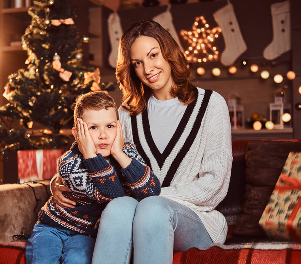 Mãe abraçando seu menino bonito enquanto sentado no sofá no quarto decorado durante o Natal
. - Foto, Imagem