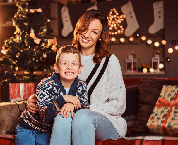Mom hugging her cute little boy while sitting on sofa in decorated room during Christmas time. - Zdjęcie, obraz