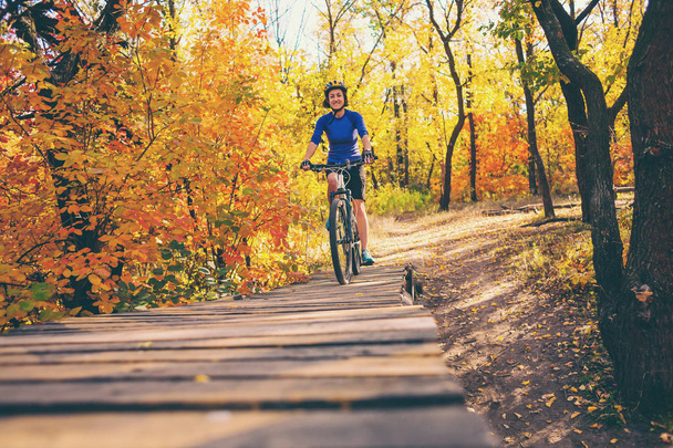 La chica conduce en un trampolín de madera. Mujer montando su bicicleta en el bosque. Preparándose para saltar. Un hobby extremo. Ciclista en el parque de otoño
. - Foto, imagen