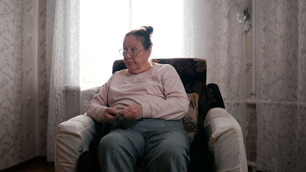 Μια ηλικιωμένη γυναίκα με το χόμπι της κάθεται σε μια καρέκλα και πλέξιμο στο φόντο του παραθύρου, κοιτάζοντας με περιφρόνηση - Φωτογραφία, εικόνα
