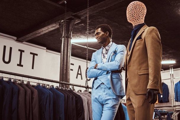 Elegantemente vestido homem afro-americano posando com braços cruzados perto de manequim em uma loja de moda masculina clássica
. - Foto, Imagem
