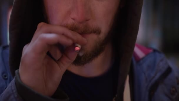 Face of young cute bearded guy dressed in hoodie is smoking on camera in street - Footage, Video