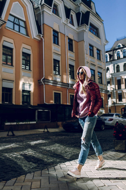 Street fashion concept - portrait of a beautiful girl sitting outside, blue jeans, sunglasses, hard lights, red jacket, autumn weather - Φωτογραφία, εικόνα