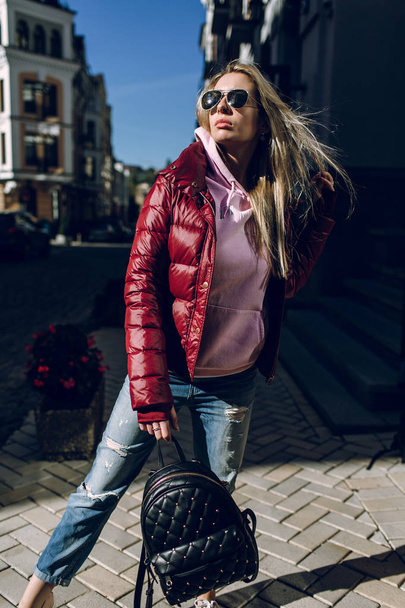 Street fashion concept - portrait of a beautiful girl sitting outside, blue jeans, sunglasses, hard lights, red jacket, autumn weather - Foto, imagen