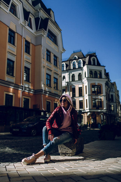 Street fashion concept - portrait of a beautiful girl sitting outside, blue jeans, sunglasses, hard lights, red jacket, autumn weather - Foto, imagen