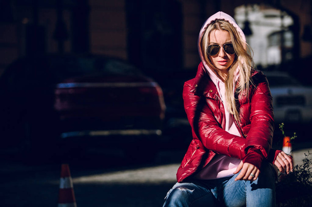 Street fashion concept - portrait of a beautiful girl sitting outside, blue jeans, sunglasses, hard lights, red jacket, autumn weather - Photo, image