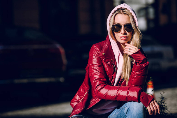 Street fashion concept - portrait of a beautiful girl sitting outside, blue jeans, sunglasses, hard lights, red jacket, autumn weather - Фото, изображение