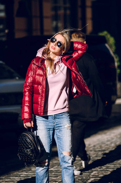 Street fashion concept - portrait of a beautiful girl sitting outside, blue jeans, sunglasses, hard lights, red jacket, autumn weather - Foto, immagini