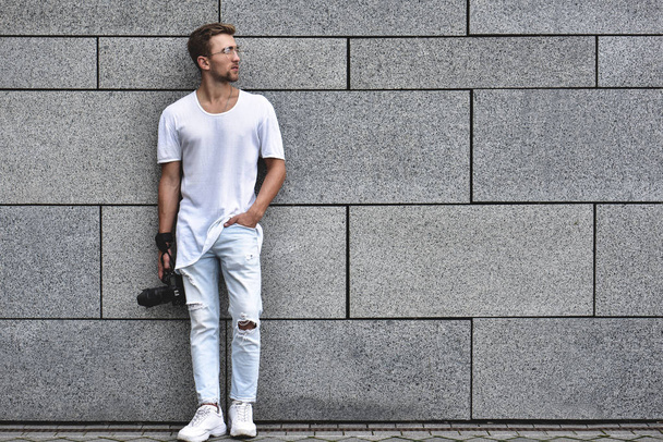 Picture of handsome Caucasian guy on grey textured wall in white T-shirt and white jeans with camera hanging on strap on his arm. - Photo, Image