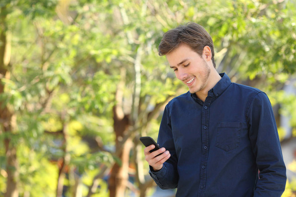 Happy man checking smart phone content walking in a park - Фото, изображение