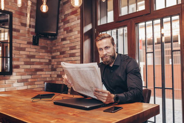 Portrait of a young bearded businessman sitting at the table and reading newspapers. - Foto, afbeelding
