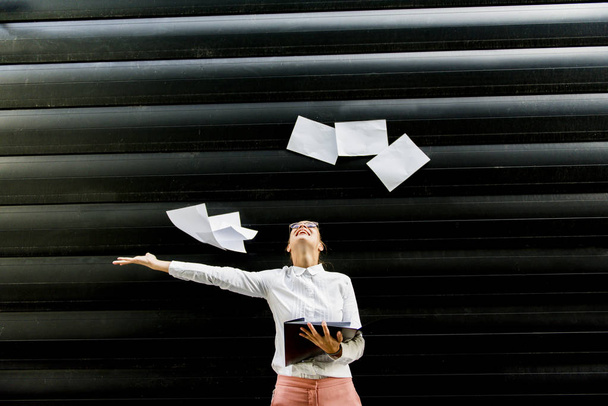 Портрет молодой предпринимательницы в деловой одежде, выбрасывающей бумажные документы на улицу
 - Фото, изображение