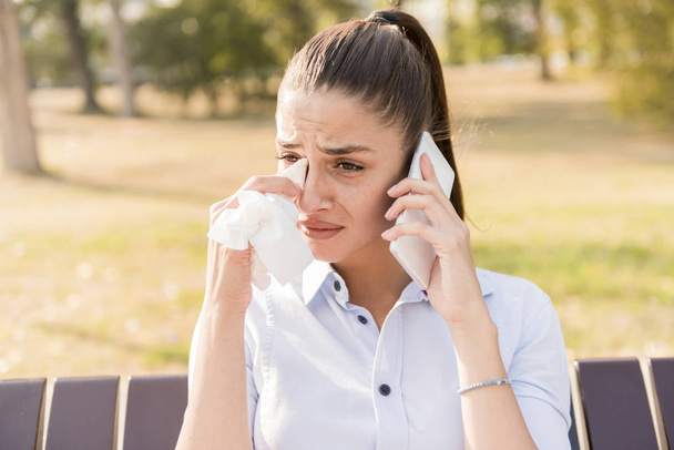 Mujer joven triste llora mientras habla en el teléfono móvil en el parque en el banco
 - Foto, Imagen