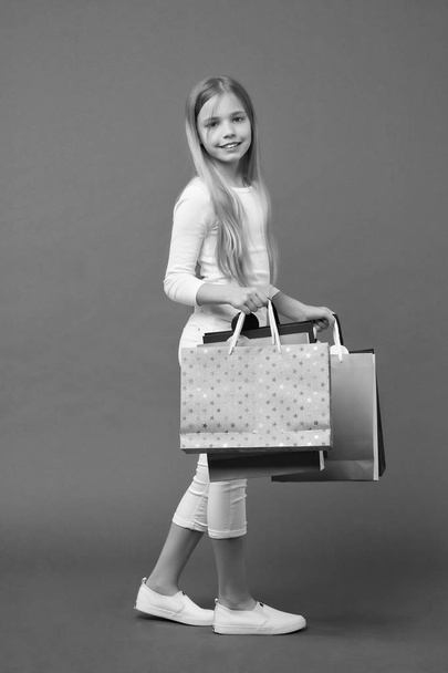 Happy kid carry shopping bags on violet background. Little shopaholic smile with paper bags. Girl shopper smiling with paperbags. Childhood. Holidays preparation and celebration. Shopping and sale - Фото, зображення