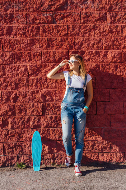 Portrait of a smiling woman dressed in overalls and sunglasses standing with her skateboard next to the red wall. - Foto, immagini