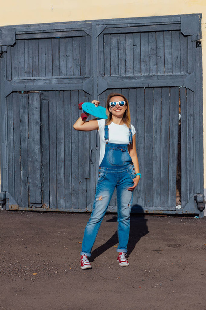 Portrait of a smiling woman dressed in overalls and sunglasses standing with her skateboard next to the old wooden wall. - Photo, Image