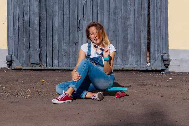 Portrait of a smiling woman dressed in overalls and sunglasses sitting on her skateboard next to the old wooden wall. - Photo, Image