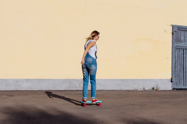 Portrait of a smiling woman dressed in overalls and sunglasses riding on her plastic skateboard next to the old wooden wall. - Фото, изображение