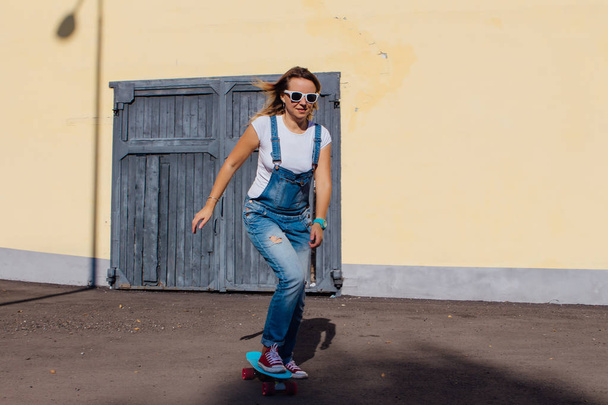 Portrait of a smiling woman dressed in overalls and sunglasses riding on her plastic skateboard next to the old wooden wall. - Photo, Image