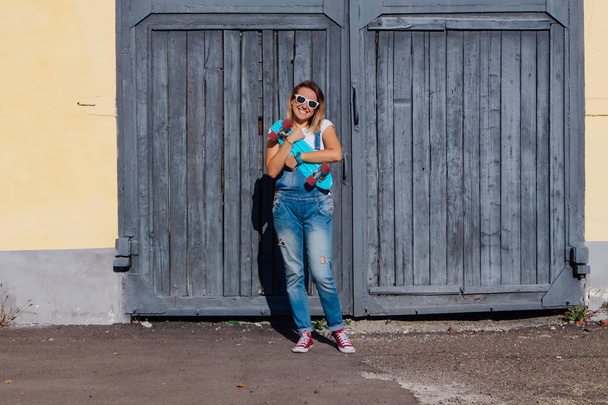 Portrait of a smiling woman dressed in overalls and sunglasses standing with her skateboard next to the old wooden wall. - Foto, Imagen