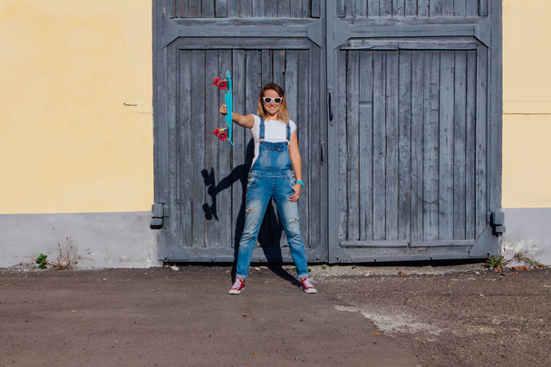Портрет усміхненої жінки, одягненої в комбінезон і сонцезахисні окуляри, що стоять зі скейтбордом поруч зі старою дерев'яною стіною
. - Фото, зображення