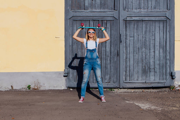 Portrait of a smiling woman dressed in overalls and sunglasses standing with her skateboard next to the old wooden wall. - Zdjęcie, obraz