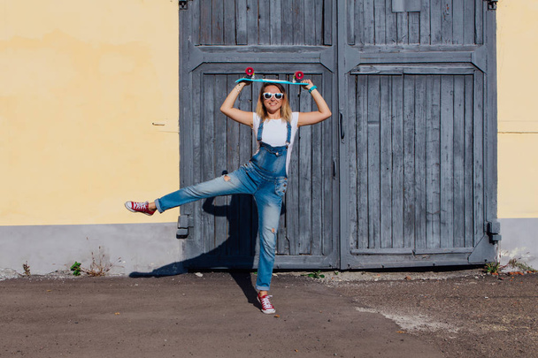 Portrait of a smiling woman dressed in overalls and sunglasses standing with her skateboard next to the old wooden wall. - Фото, изображение