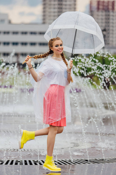 透明傘と黄色のブーツでお下げ髪若いきれいな女の子は噴水のそばに立ちます。雨の日の市. - 写真・画像