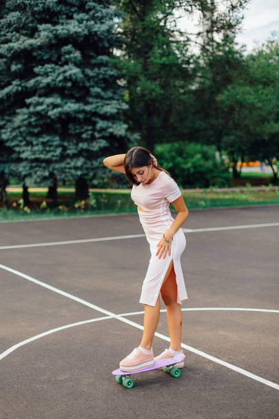 Portrait of a smiling charming brunette female standing on her skateboard on a basketball court. Happy woman with trendy look taking break during sunset. - Фото, зображення