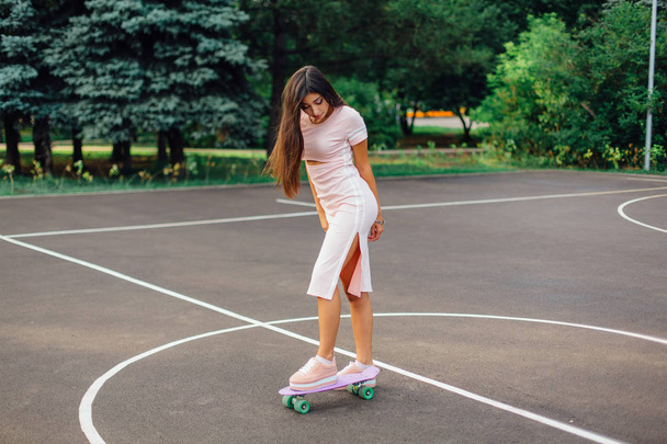 Portrait of a smiling charming brunette female standing on her skateboard on a basketball court. Happy woman with trendy look taking break during sunset. - Photo, Image
