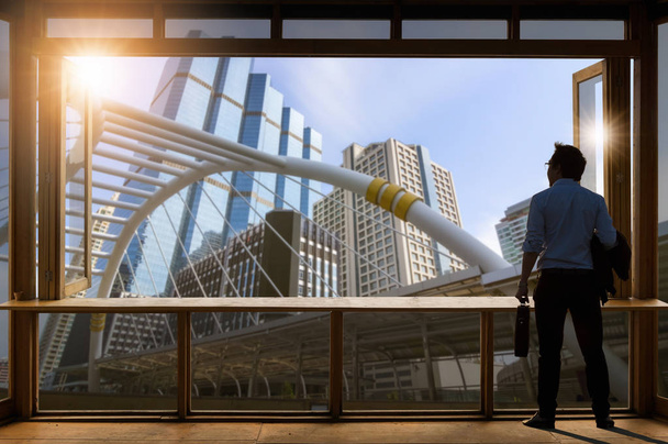 Konzeptvision, junger Geschäftsmann in bequemer Freizeitjacke steht im Arbeitszimmer am Glasfenster und schaut durch die modernen Gebäude in der Skyline. - Foto, Bild