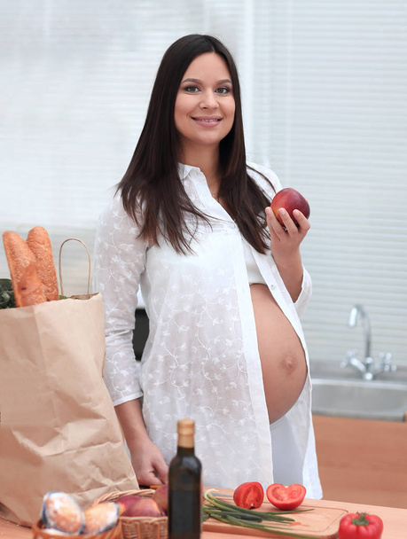 young pregnant woman holding an Apple,standing in the kitchen - Photo, image