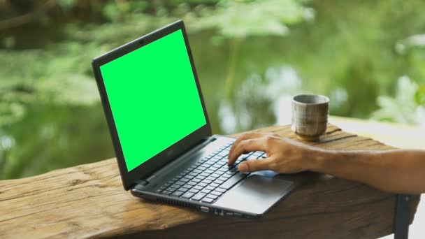 Рука людини використовує ноутбук з зеленим екраном на дерев'яному столі з чашкою напою біля ставка вранці
. - Кадри, відео