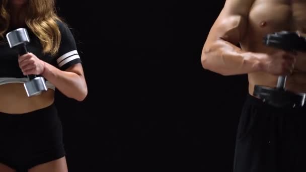 Hombre y mujer atléticos flexiona sus manos con mancuernas, entrenando sus bíceps sobre un fondo negro en el estudio. Se encuentran en diferentes bordes del marco. Centro de texto
 - Metraje, vídeo