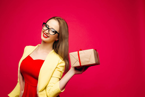 Beautiful happy woman in fashionable cat-eye glasses in bright dress holding Christmas present. - Foto, imagen