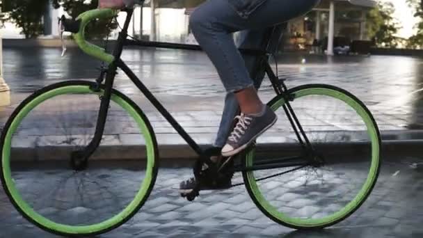 Nahaufnahmen von weiblichen Füßen, die morgens mit dem Fahrrad durch eine gepflasterte Stadtstraße mit nassem Asphalt fahren. Seitenansicht einer jungen Frau auf einem Trekkingrad mit grünen Rädern, die Turnschuhe und Jeans trägt - Filmmaterial, Video