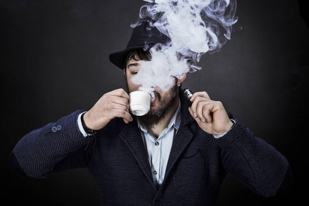 bärtiger Typ mit Hut trinkt Kaffee und raucht eine elektronische Zigarre auf schwarzem Hintergrund - Foto, Bild