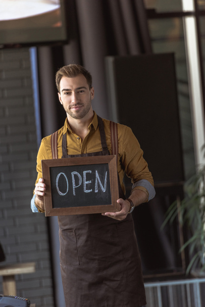 handsome waiter in apron holding chalkboard with open inscription in cafe - Photo, Image