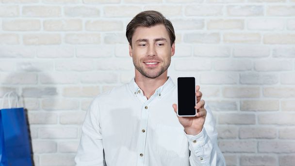Lächelnder junger Mann mit Smartphone und leerem Bildschirm bei der Arbeit im Geschäft - Foto, Bild