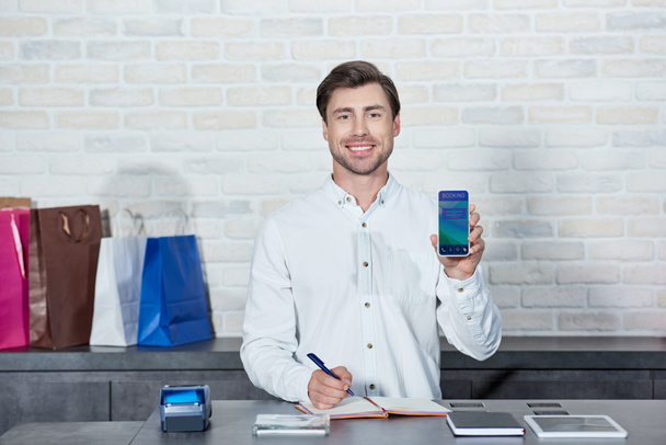 handsome male seller holding smartphone with booking application and smiling at camera in store  - Foto, Bild