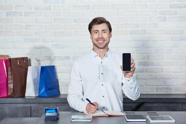 handsome young salesman holding smartphone with blank screen and smiling at camera in shop - Photo, image