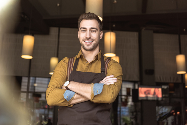 handsome young waiter with crossed arms looking at camera in cafe - Foto, Imagen