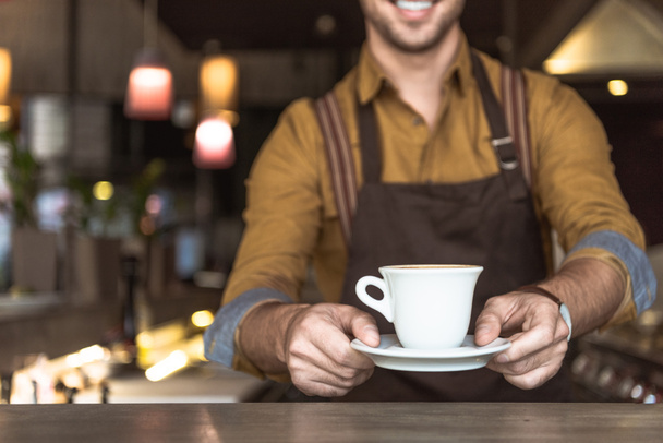 обрізаний знімок усміхненого молодого бариста, що тримає чашку кави
 - Фото, зображення