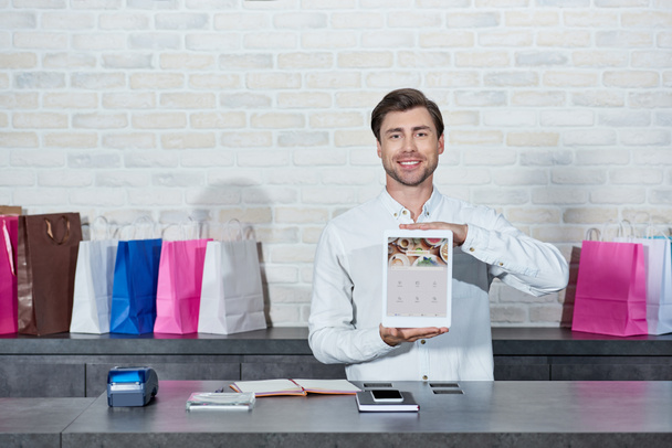 handsome young salesman holding digital tablet with foursquare application and smiling at camera in shop   - Fotoğraf, Görsel