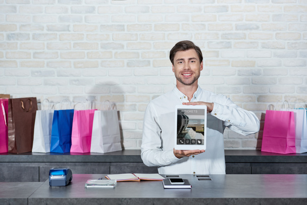 przystojny młody sprzedawca gospodarstwa cyfrowy tablet z aplikacją bilety i uśmiecha się do kamery w sklepie - Zdjęcie, obraz