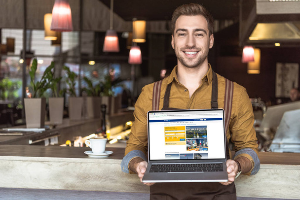 красивий молодий офіціант тримає ноутбук з бронювання сайт на екрані в кафе - Фото, зображення