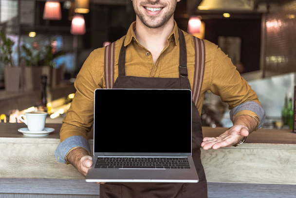 colpo ritagliato di giovane cameriere sorridente che tiene il computer portatile con schermo vuoto in caffè
 - Foto, immagini