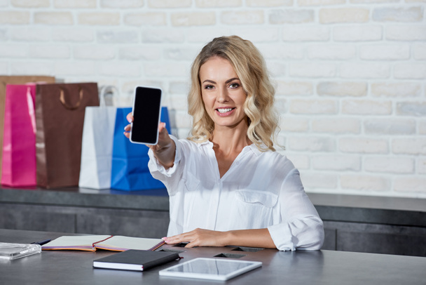 beautiful young seller holding smartphone with blank screen and smiling at camera in shop  - Photo, Image