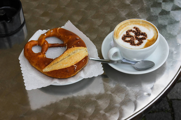 italian cappuccino with german pretzel with chocolate artwork on creme of coffee on metal table top in south german historical city near munich and stuttgart - Photo, Image